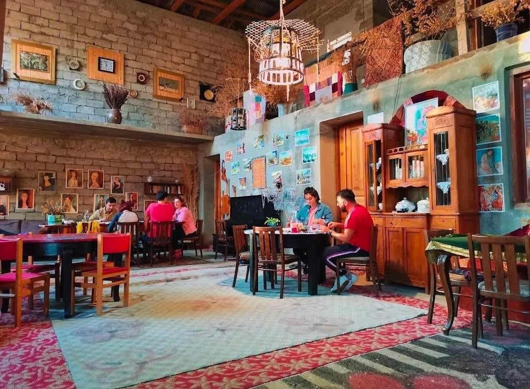 Colorful living room set with multiple people sitting at tables, exposed brick walls, wooden furniture, and vibrant decorations throughout